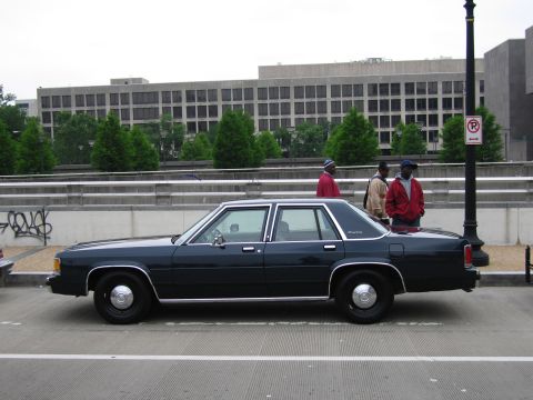 1991 Ford Crown Victoria - A dark 1991 Ford Crown Victoria sedan is seen from the side, with three men standing near the rear.