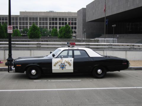 1991 Ford Crown Victoria - A side view of a vintage highway patrol car parked on a street in front of a large building.