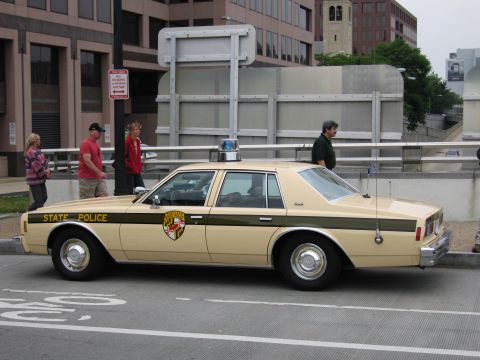 1991 Ford Crown Victoria - A tan state police car is parked on the street, with people walking nearby.