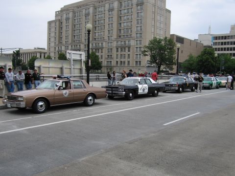 1991 Ford Crown Victoria - A convoy of older model police vehicles is parked on a city street.