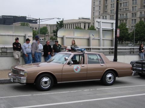 1991 Ford Crown Victoria - A brown police car with a light bar is parked on the street, with a group of people standing next to it.