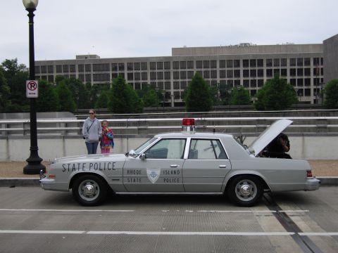 1991 Ford Crown Victoria - A gray Rhode Island State Police car is parked on a street with two people standing nearby and the trunk open.