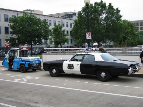1991 Ford Crown Victoria - Two police vehicles, an electric cart and a sedan, are parked side-by-side on a city street.