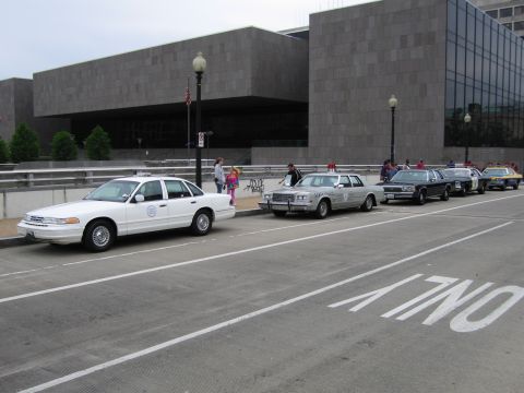 1991 Ford Crown Victoria - A line of police cars is parked on the street in front of a modern building.