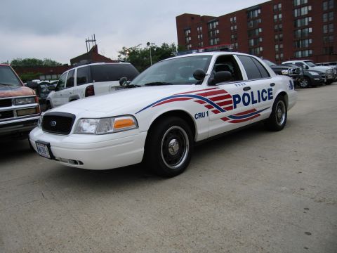 1991 Ford Crown Victoria - A white police car is parked on a concrete surface with other vehicles and buildings in the background.