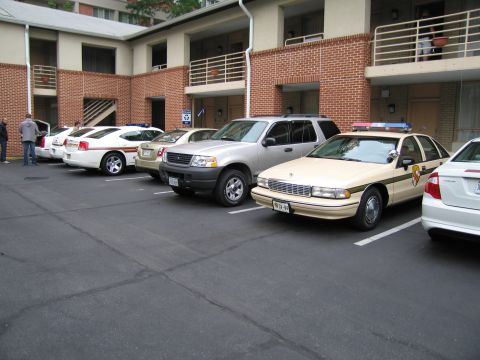 1991 Ford Crown Victoria - A line of vehicles, including police cruisers and an SUV, are parked in a lot outside a multi-story building.