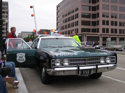 1991 Ford Crown Victoria - A vintage police car, likely from the late 1960s, is parked on a street with people around it.