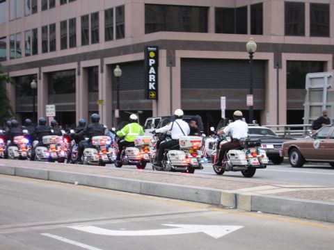 1991 Ford Crown Victoria - A line of police motorcycles rides down a city street, with officers in uniform and a building in the background.
