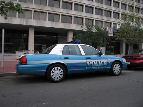 1991 Ford Crown Victoria - A blue and white police car is parked on the street in front of a building, with another car visible to its right.
