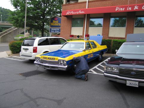 1991 Ford Crown Victoria - A person works on the front wheel of a vintage New York State Police car parked next to a van and another car.