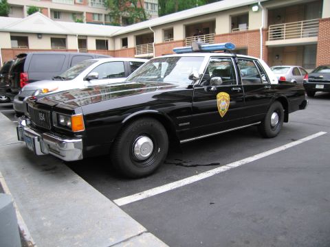 1991 Ford Crown Victoria - A police car with a light bar and official markings is parked in a lot next to other vehicles.