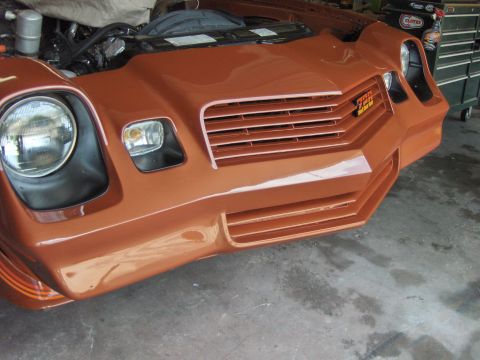 1980 Chevrolet Camaro - The front end of an orange sports car is shown, with the hood open, revealing engine parts.