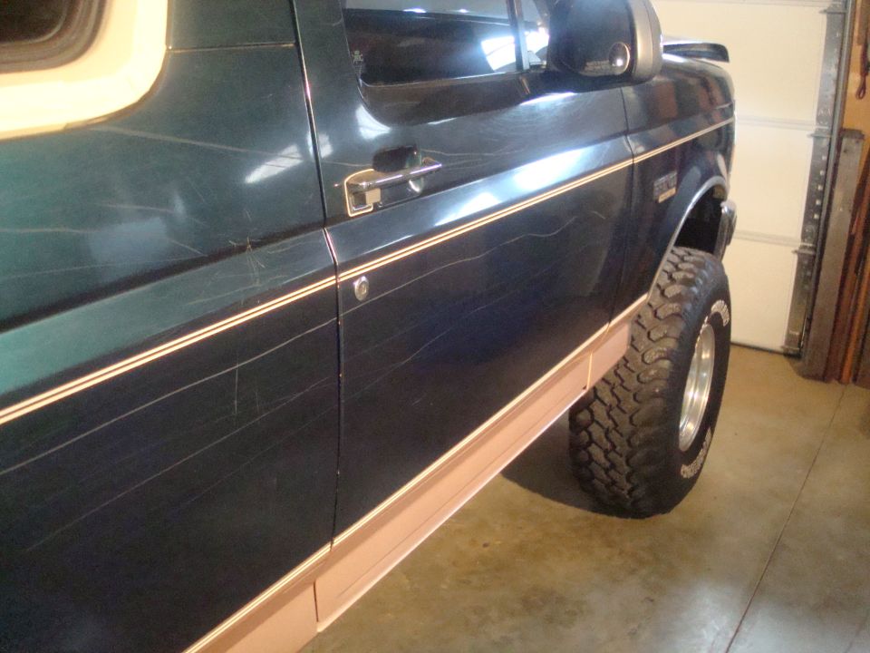 1995 Ford Bronco side door with chrome handle and pinstriping, showing heavy scratches. Lifted suspension with off-road tire visible