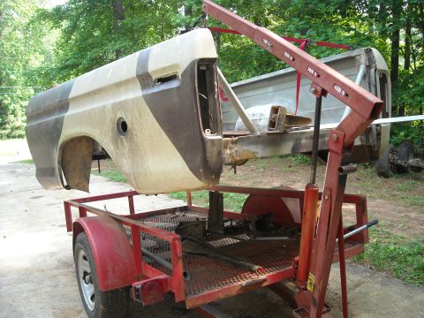 1979 Ford F-150 - A truck bed section, painted in a camouflage pattern, is suspended by an engine hoist over a trailer.