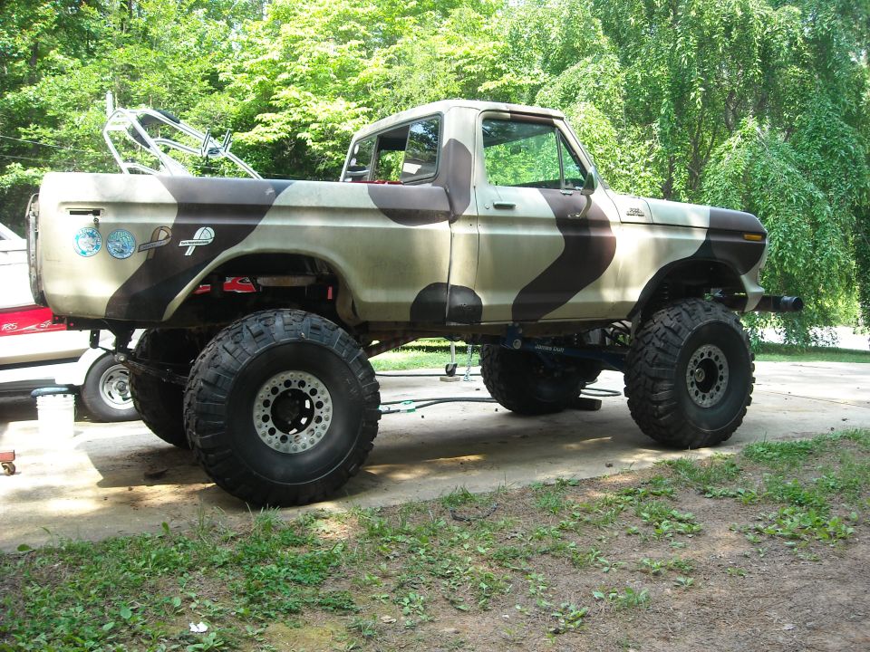 Lifted pickup truck with camouflage paint, large off-road tires, and a boat in the background