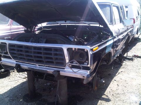 1986 Ford Bronco - This black SUV, likely from the early 1980s, sits in a junkyard with its hood open and missing headlights.