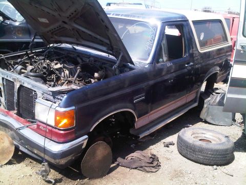 1986 Ford Bronco - Junkyard Bronco missing wheels and parts, hood is open showing the engine bay.