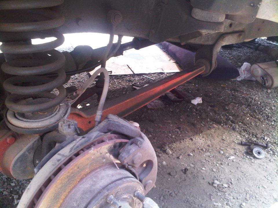 Close-up of a vehicle's rear suspension, showing the coil spring, brake rotor, and control arm. The vehicle appears to be on uneven ground, possibly a junkyard
