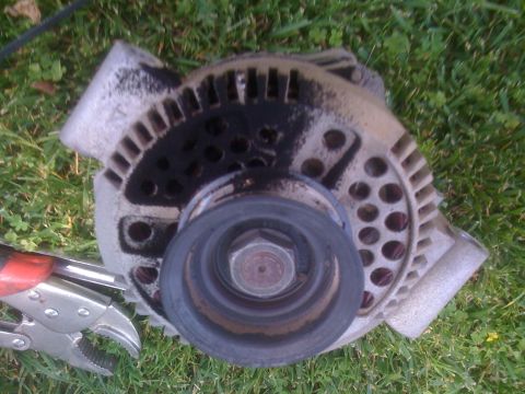 1996 Ford C-350 (Centurion Conversions) - A removed automotive alternator lying pulley-up in green grass, its cast-aluminum housing streaked with black grease and the vented end cov…