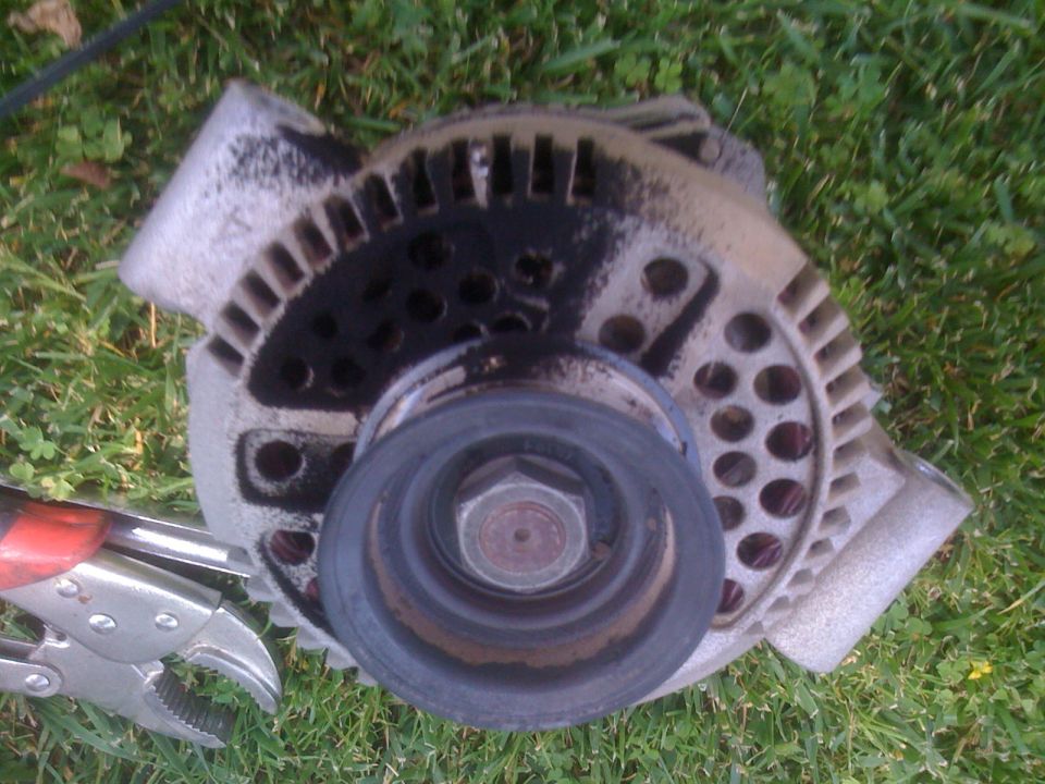 Greasy removed alternator lying pulley-up in grass beside red-handled locking pliers