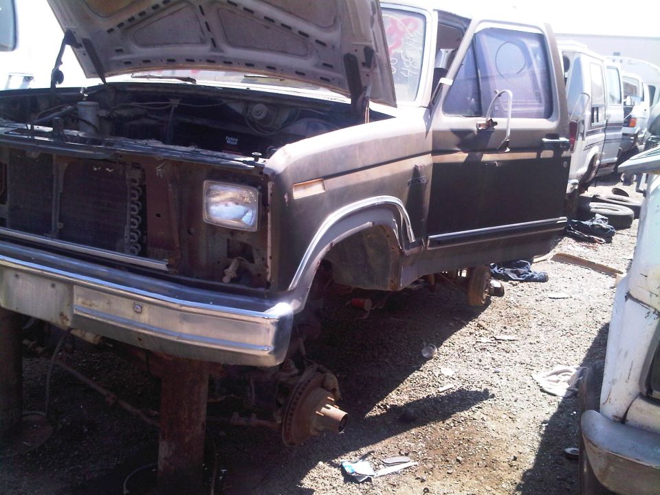 Brown Ford truck with hood and door open, missing front wheels, in a junkyard