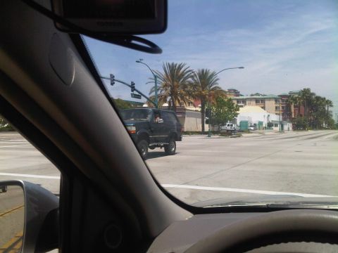 1986 Ford Bronco - A dark SUV is seen driving through an intersection on a sunny day, with palm trees and buildings lining the street.