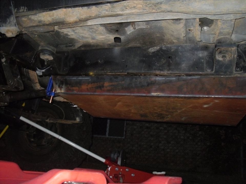 Underbody armor plating being installed on a vehicle, with a jack and gas can visible
