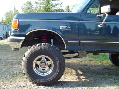 1993 Ford Bronco - Close side view of a lifted dark green Ford Bronco's front corner on a grass lot, showing a red coil-spring shock at the front axle, radius…