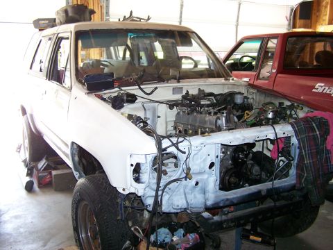 1988 Suzuki Samurai - White SUV undergoing engine work in a garage, with a red pickup truck parked nearby.