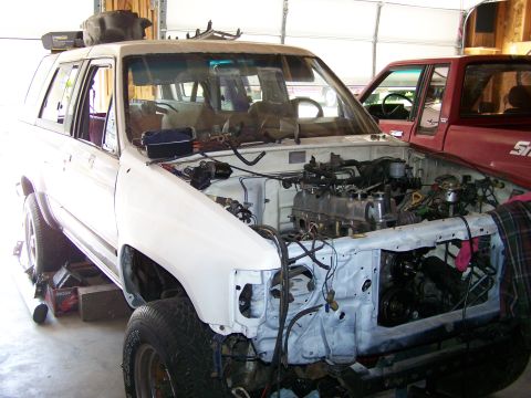 1988 Suzuki Samurai - A white SUV is in a garage with its engine compartment open, revealing the engine bay. A red truck is parked nearby.
