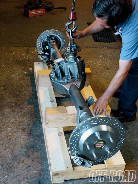 1990 Ford Bronco - A rear axle assembly with drilled rotors is prepared for installation, lifted by a crane in a workshop.