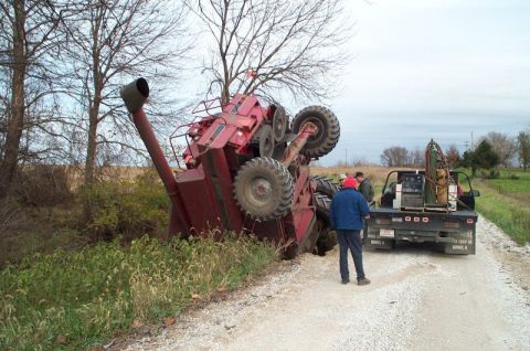 1994 Ford Bronco - A red combine harvester has overturned on its side in a ditch, with a pickup truck and onlookers present on the adjacent dirt road.