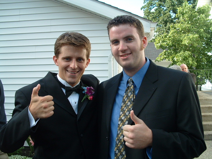 Two people in tuxedo and suit giving thumbs-up outside a white building