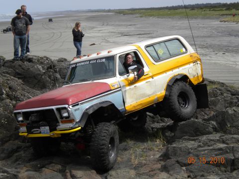 1994 Ford Explorer - A vintage Ford Bronco navigates a rocky terrain on a beach, with onlookers in the background.