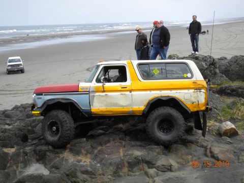 1994 Ford Explorer - Off-road modified Ford Bronco navigating rocky terrain on a beach, with another vehicle visible in the distance.