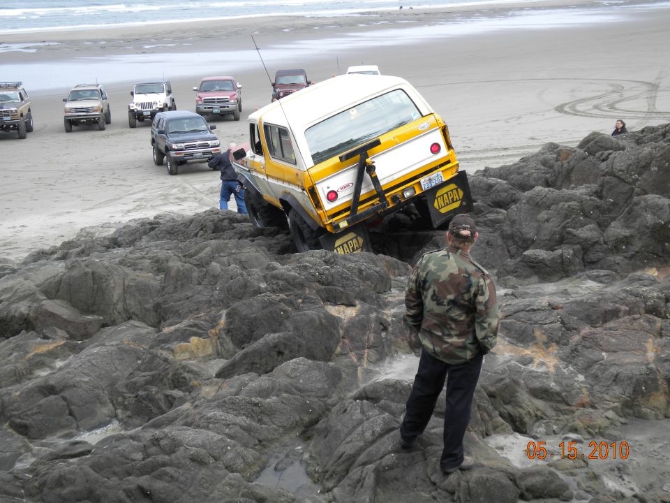 Yellow and white SUV stuck on rocks by the ocean, with other vehicles on the beach behind it. Someone is trying to push it