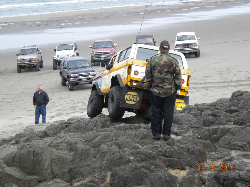 Yellow and white off-road vehicle with NAPA decals on a rocky beach, other SUVs and trucks in background