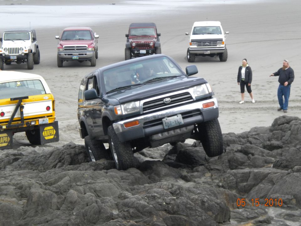 Dark Toyota 4Runner navigating rocky terrain on a beach, with other vehicles and people in the background