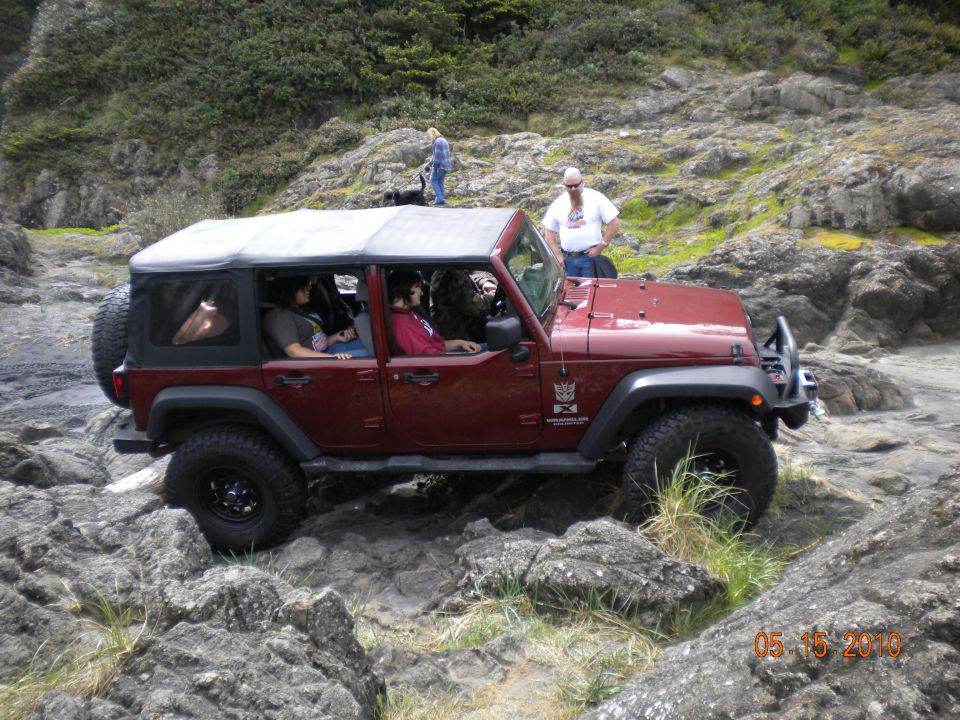 Red Jeep Wrangler with soft top driving over large rocks near a body of water. People are inside the vehicle and standing nearby