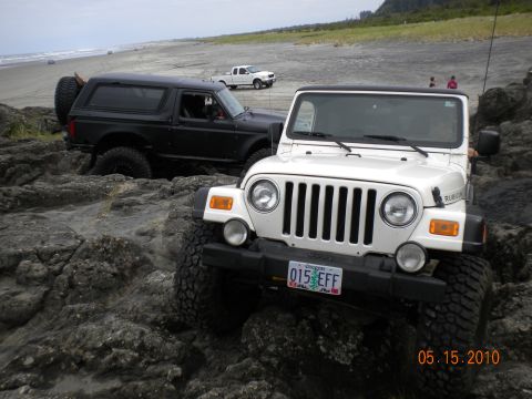 1994 Ford Explorer - Two off-road vehicles, a white Jeep Wrangler and a black Ford Bronco, are parked on a rocky shore with a white pickup truck in the backgrou…