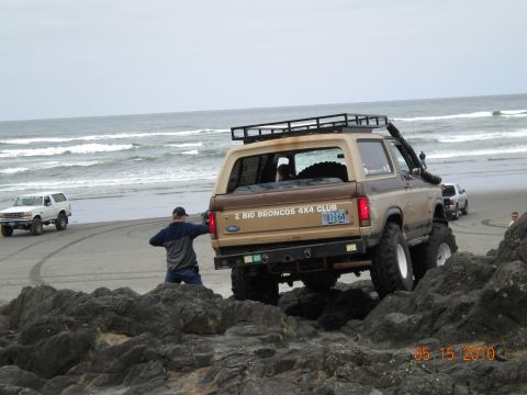 1994 Ford Explorer - A tan Ford Bronco II navigates rocky terrain on a beach, with another white Bronco visible in the background.