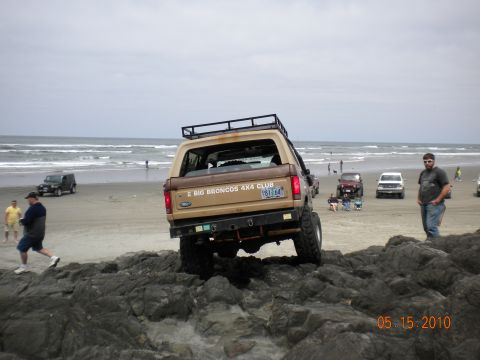 1994 Ford Explorer - A lifted tan Ford Bronco navigates rocky terrain on a beach, with other vehicles and people in the background.