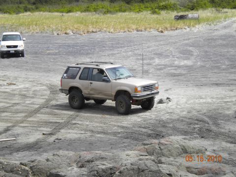 1994 Ford Explorer - A tan 1994 Ford Explorer with lifted suspension and off-road tires drives on a sandy beach, with a white SUV in the distance.