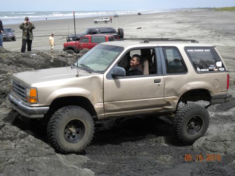 1994 Ford Explorer - A woman drives a lifted 1994 Ford Explorer over rocks on a beach, with other vehicles visible in the background.