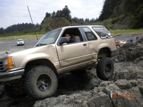 1994 Ford Explorer - A gold 1994 Ford Explorer 3-door navigates rocky terrain on a beach, with a white SUV visible in the distance.