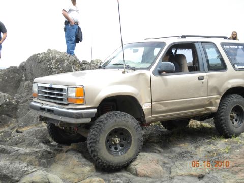 1994 Ford Explorer - A gold Ford Explorer navigates a rocky terrain, with two people standing on rocks in the background.