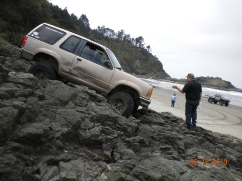 1994 Ford Explorer - A tan SUV navigates rocky terrain near a beach, with another vehicle visible on the sand.