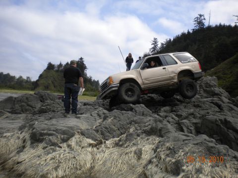 1994 Ford Explorer - Tan 2-door SUV with off-road tires navigates a rocky terrain on a beach.