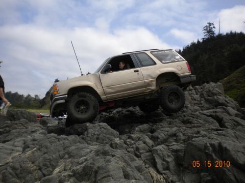 1994 Ford Explorer - A tan 1994 Ford Explorer navigates a rocky coastal terrain, its lifted suspension and off-road tires.