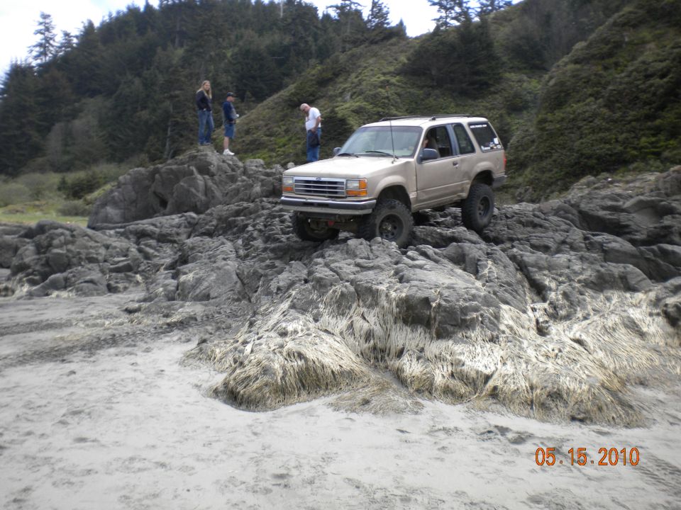 Tan lifted two-door Ford Explorer rock crawling on coastal rocks with people watching
